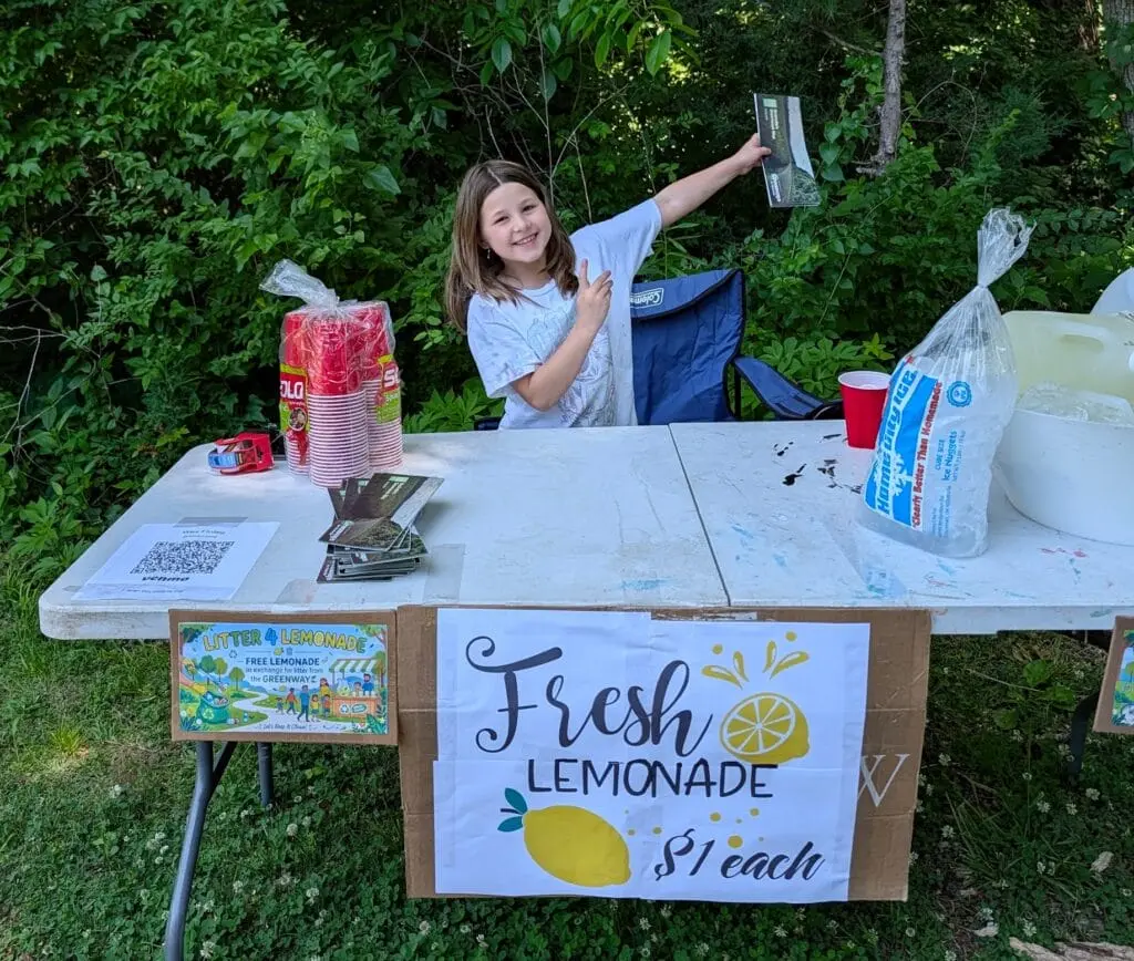 Audrey smiling behind her homemade lemonade stand with red cups, ice, and a hand-lettered sign