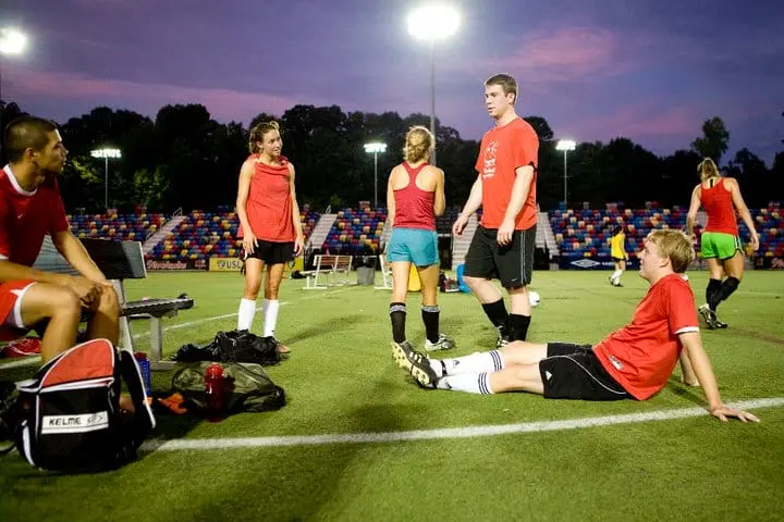 Adult recreational soccer players resting on a sideline under stadium lights at dusk